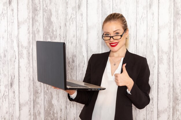 Vue de face jeune femme d'affaires dans des vêtements stricts veste noire à l'aide de son ordinateur portable souriant sur un bureau blanc