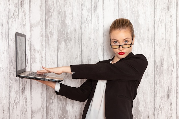 Vue de face jeune femme d'affaires dans des vêtements stricts veste noire à l'aide de son ordinateur portable sur une surface blanche