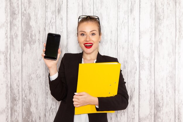 Vue de face jeune femme d'affaires dans des vêtements stricts veste noire tenant des documents et téléphone sur une surface blanche