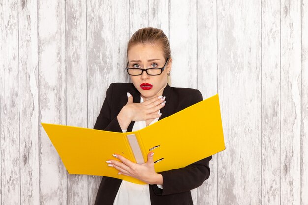 Vue de face jeune femme d'affaires dans des vêtements stricts veste noire tenant des fichiers et des documents sur un bureau blanc clair