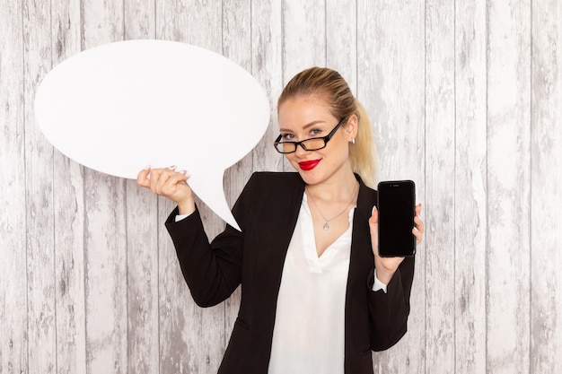 Vue de face jeune femme d'affaires dans des vêtements stricts veste noire tenant son téléphone et panneau blanc sur une surface blanche