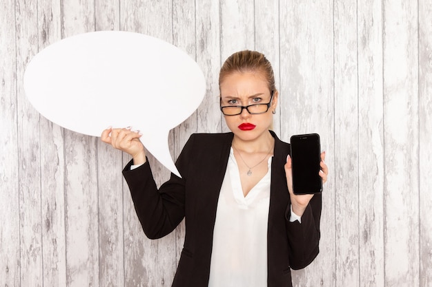 Vue de face jeune femme d'affaires dans des vêtements stricts veste noire tenant son téléphone et panneau blanc sur une surface blanche