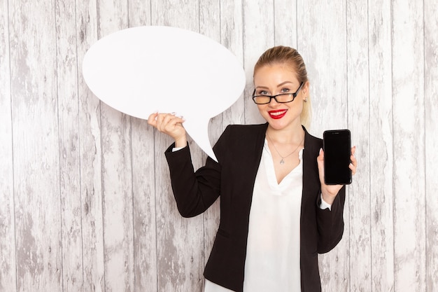 Photo gratuite vue de face jeune femme d'affaires dans des vêtements stricts veste noire tenant son téléphone et panneau blanc sur une surface blanche