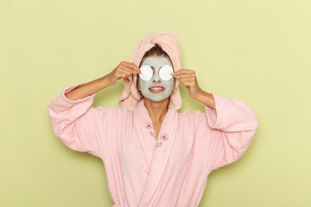 Vue de face jeune femme après la douche en peignoir rose couvrant ses yeux avec du coton sur une surface verte