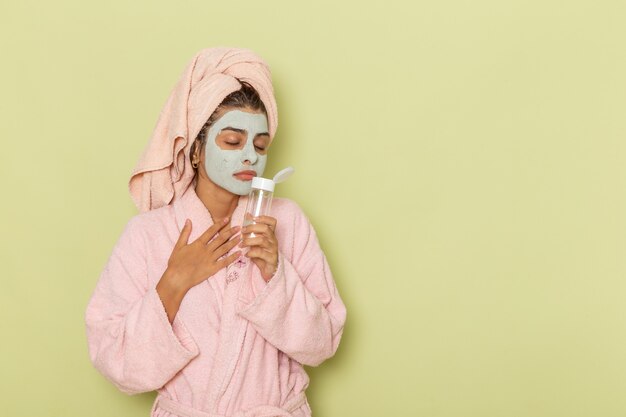 Vue de face jeune femme après la douche en peignoir rose tenant un démaquillant sur un bureau vert