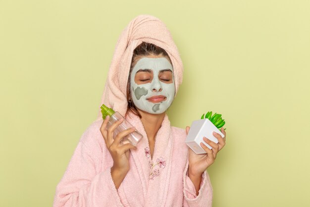 Vue de face jeune femme après la douche en peignoir rose tenant des démaquillants sur le sol vert crème masque douche selfcare bain de beauté