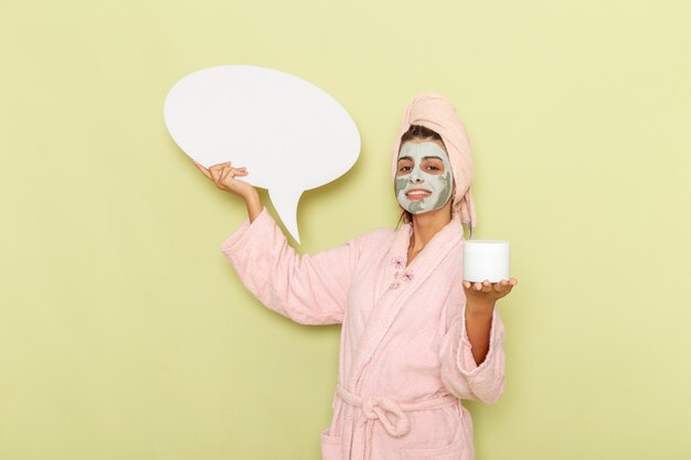 Vue de face jeune femme après la douche en peignoir rose tenant une pancarte blanche et crème sur surface verte