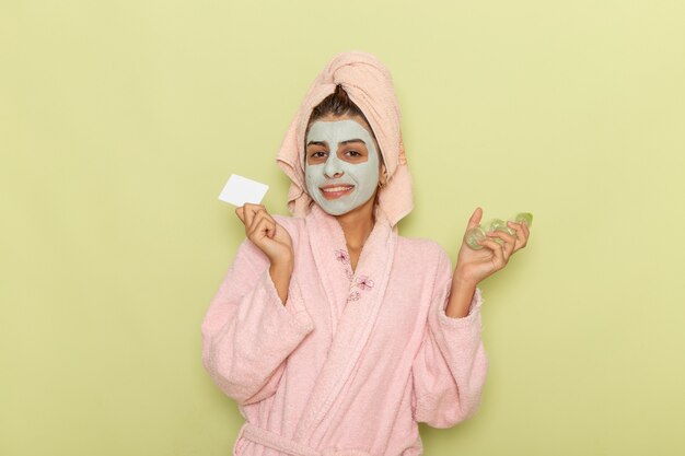 Vue de face jeune femme après la douche en peignoir rose tenant des sprays et une carte sur un bureau vert