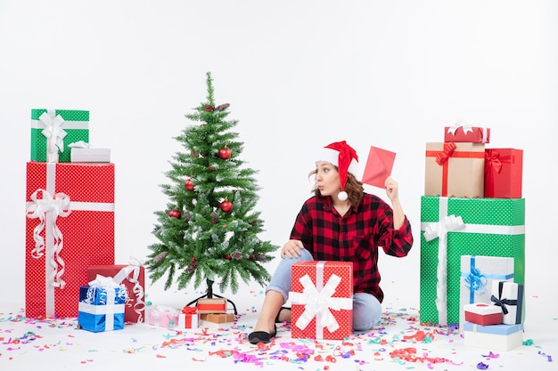 Vue de face de la jeune femme assise autour de cadeaux holding enveloppe sur mur blanc