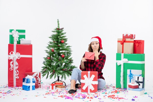 Vue de face de la jeune femme assise autour de cadeaux holding enveloppe sur mur blanc