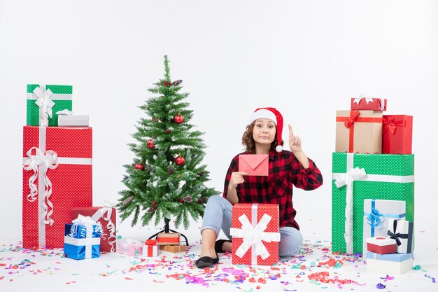 Vue de face de la jeune femme assise autour de cadeaux holding enveloppe sur mur blanc