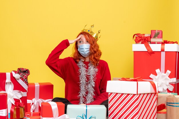 Vue de face de la jeune femme assise autour de cadeaux en masque stérile sur mur jaune