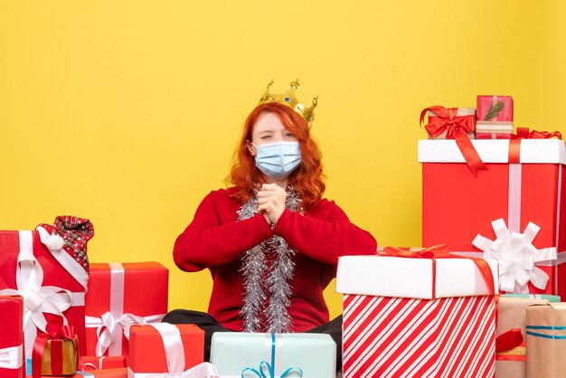 Vue de face de la jeune femme assise autour de cadeaux en masque stérile sur mur jaune