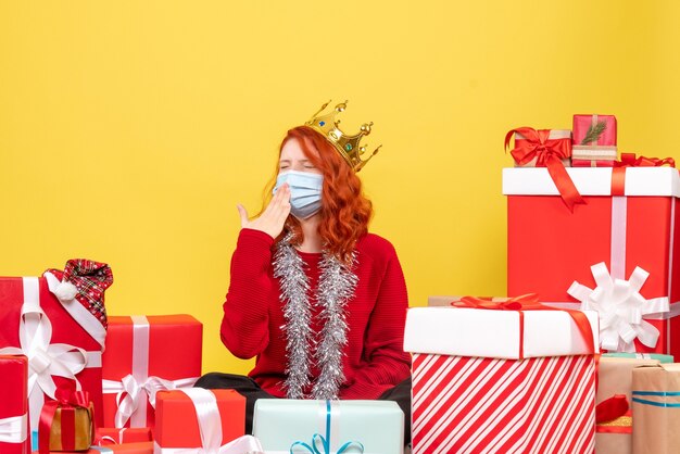 Vue de face de la jeune femme assise autour de cadeaux en masque stérile sur mur jaune