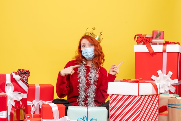 Vue de face de la jeune femme assise autour de cadeaux en masque stérile sur mur jaune