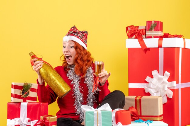 Vue de face de la jeune femme assise autour des cadeaux de Noël et célébrer avec du champagne sur le mur jaune