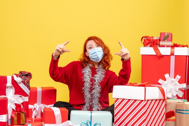 Vue de face de la jeune femme assise autour de cadeaux de Noël en masque sur mur jaune