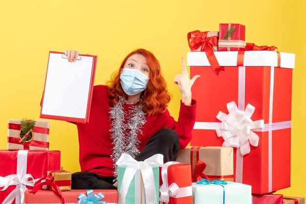 Vue de face de la jeune femme assise autour des cadeaux de Noël en masque sur un mur jaune