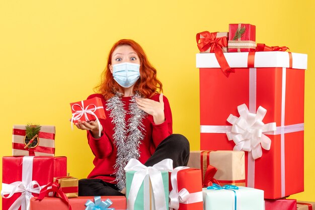 Vue de face de la jeune femme assise autour des cadeaux de Noël en masque sur un mur jaune