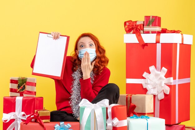 Vue de face de la jeune femme assise autour des cadeaux de Noël en masque sur un mur jaune