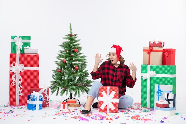 Vue de face de la jeune femme assise autour des cadeaux de Noël et petit arbre de vacances sur le mur blanc