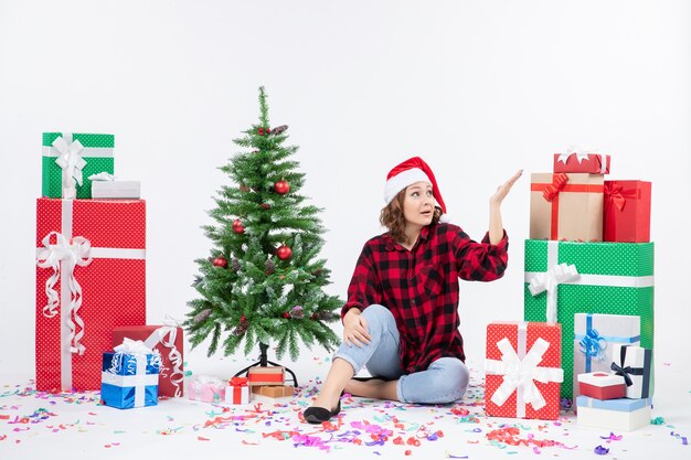 Vue de face de la jeune femme assise autour des cadeaux de Noël et petit arbre de vacances sur le mur blanc