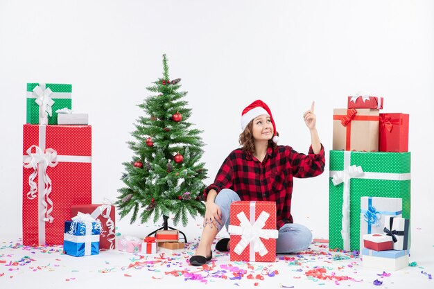 Vue de face de la jeune femme assise autour des cadeaux de Noël et petit arbre de vacances sur le mur blanc