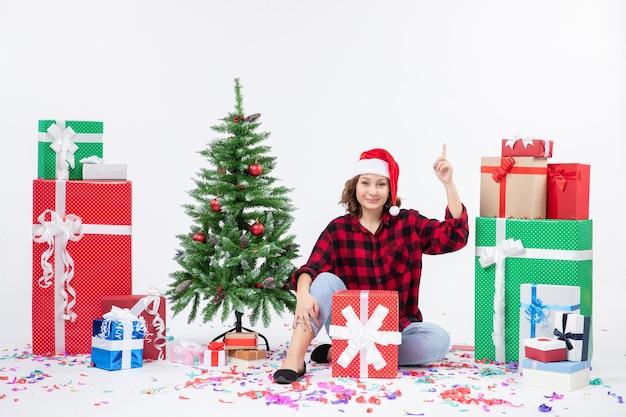 Vue de face de la jeune femme assise autour des cadeaux de Noël et petit arbre de vacances sur le mur blanc