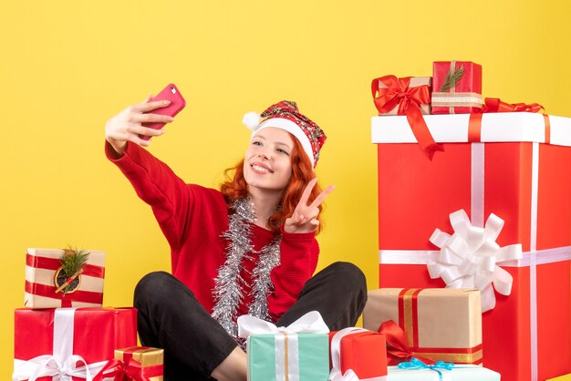 Vue de face de la jeune femme assise autour de cadeaux de Noël prenant selfie sur mur jaune