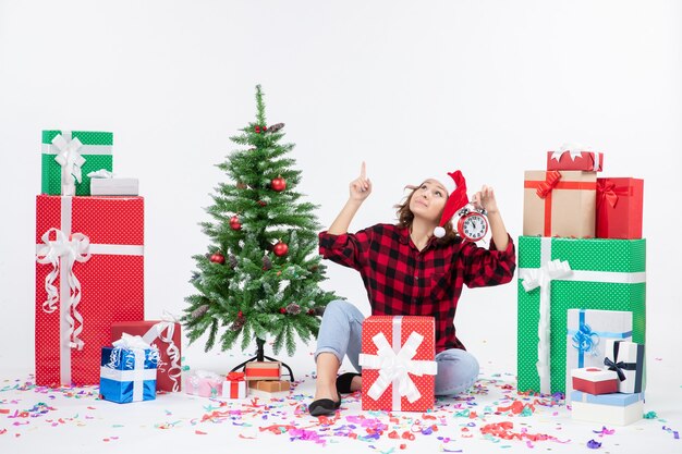 Vue de face de la jeune femme assise autour de cadeaux de Noël tenant des horloges sur un mur blanc