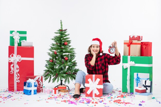 Vue de face de la jeune femme assise autour de cadeaux de Noël tenant des horloges sur un mur blanc