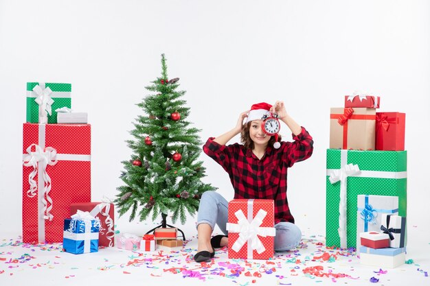 Vue de face de la jeune femme assise autour de cadeaux de Noël tenant des horloges sur un mur blanc