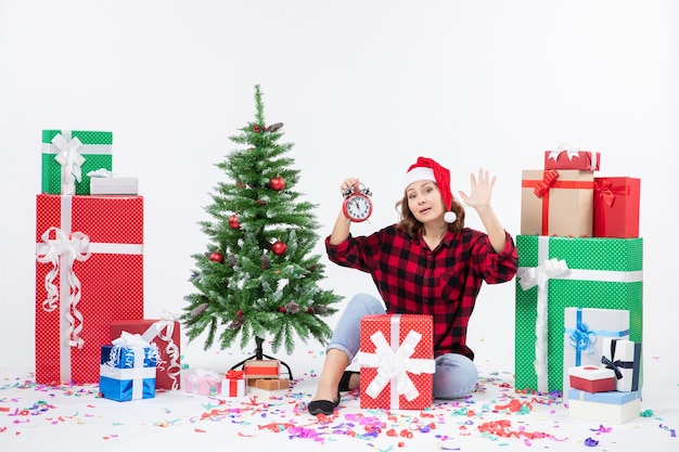 Vue de face de la jeune femme assise autour de cadeaux de Noël tenant des horloges sur un mur blanc