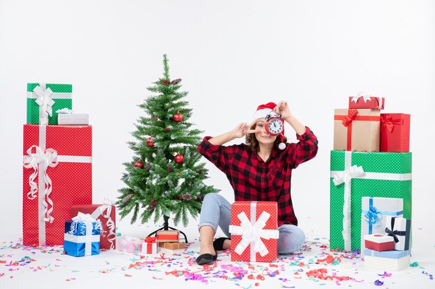 Vue de face de la jeune femme assise autour de cadeaux de Noël tenant des horloges sur un mur blanc