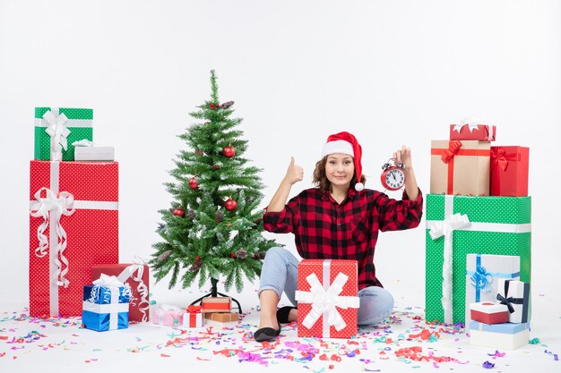 Vue de face de la jeune femme assise autour de cadeaux de Noël tenant des horloges sur un mur blanc