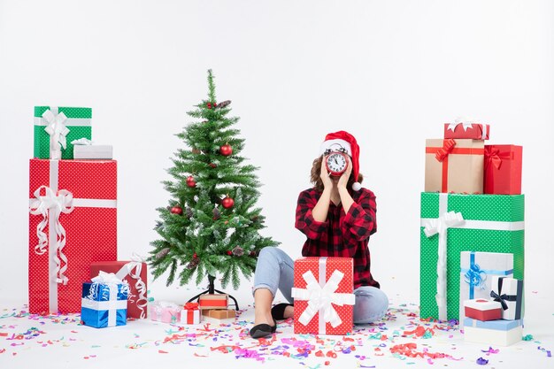 Vue de face de la jeune femme assise autour de cadeaux de Noël tenant des horloges sur un mur blanc