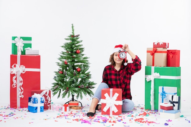 Vue de face de la jeune femme assise autour de cadeaux de Noël tenant des horloges sur un mur blanc