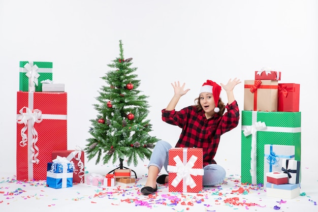 Vue de face de la jeune femme assise autour de cadeaux et petit arbre de vacances sur un mur blanc