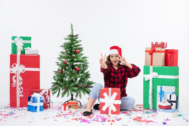 Vue de face de la jeune femme assise autour de cadeaux et petit arbre de vacances sur un mur blanc
