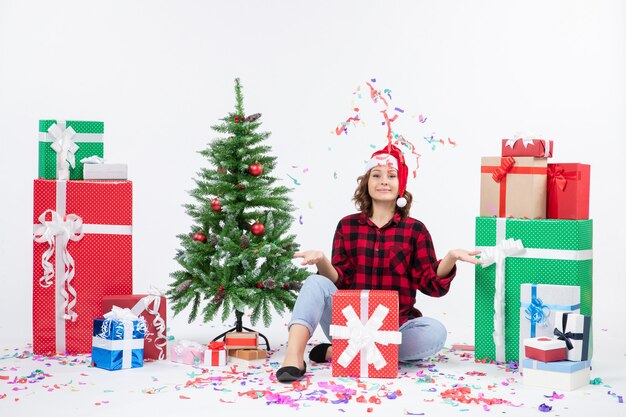 Vue de face de la jeune femme assise autour de cadeaux et petit arbre de vacances sur un mur blanc