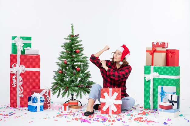 Vue de face de la jeune femme assise autour de cadeaux et petit arbre de vacances sur un mur blanc