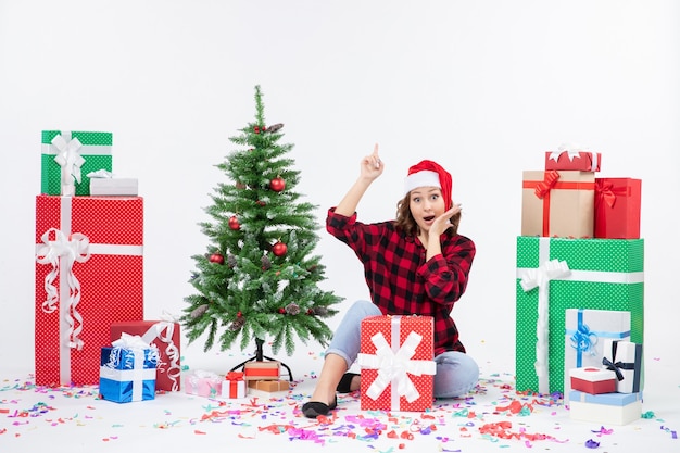 Vue de face de la jeune femme assise autour de cadeaux et petit arbre de vacances sur un mur blanc