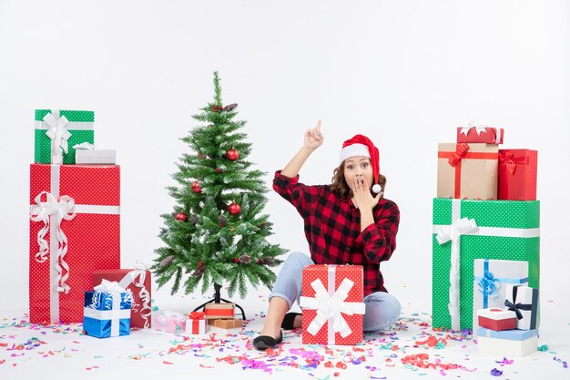 Vue de face de la jeune femme assise autour de cadeaux et petit arbre de vacances sur un mur blanc