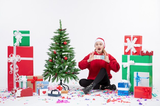 Vue de face de la jeune femme assise autour de cadeaux tenant des billets d'avion sur un mur blanc