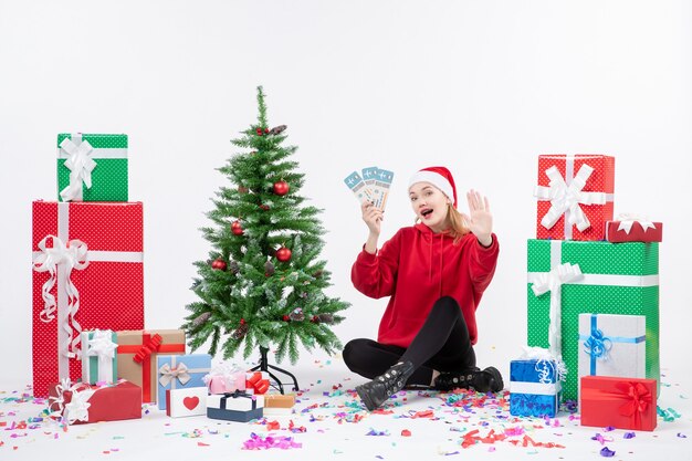 Vue de face de la jeune femme assise autour de cadeaux tenant des billets d'avion sur un mur blanc