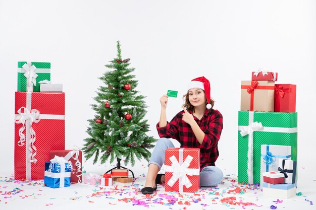 Vue de face de la jeune femme assise autour de cadeaux tenant une carte bancaire verte sur un mur blanc