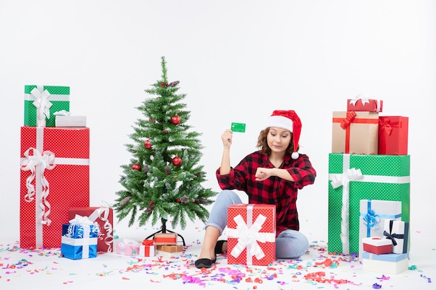 Vue de face de la jeune femme assise autour de cadeaux tenant une carte bancaire verte sur un mur blanc