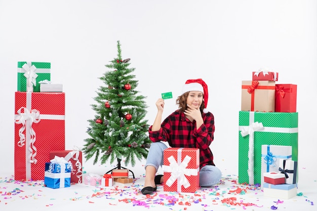 Vue de face de la jeune femme assise autour de cadeaux tenant une carte bancaire verte sur un mur blanc