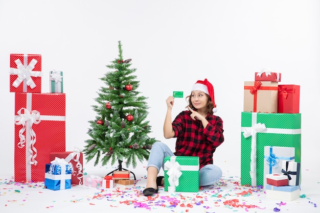 Vue de face de la jeune femme assise autour de cadeaux tenant une carte bancaire verte sur un mur blanc