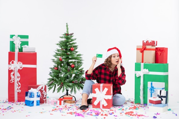 Vue de face de la jeune femme assise autour de cadeaux tenant une carte bancaire verte sur un mur blanc